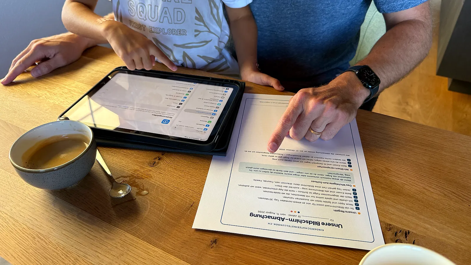 Un père et son enfant à la table : le père montre l’accord d’écran imprimé, à côté une tablette affiche les réglages ouverts